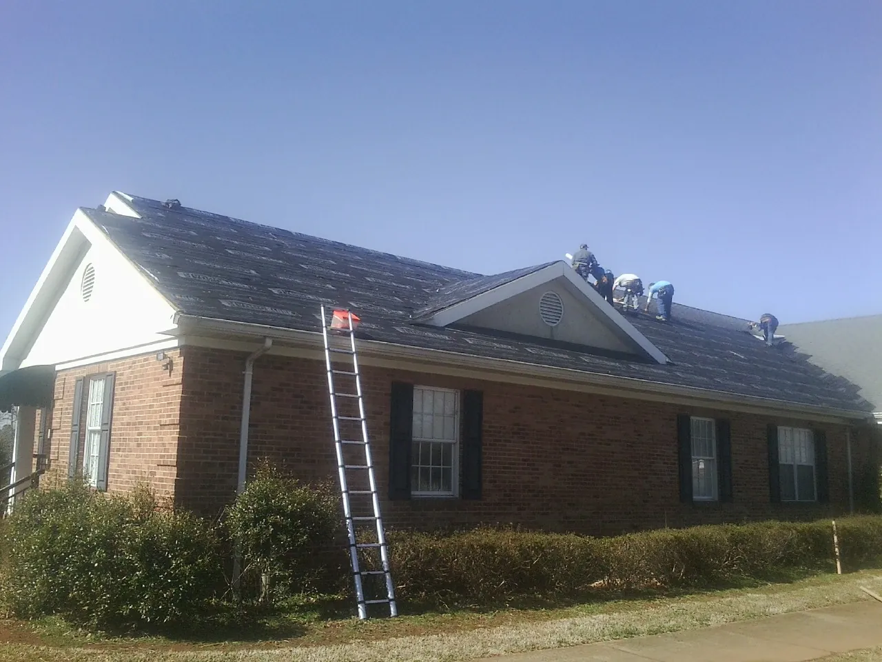 Crew tearing shingles off a brick ranch roof with a ladder set — Emergency Roof Repair work by our crew in Perrinton