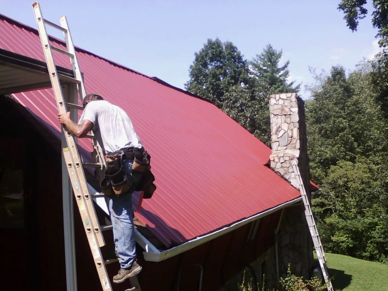 Roofer on a ladder beside a red metal roof and stone chimney — Soffit & Fascia Repair by Roofing Brothers in Perrinton